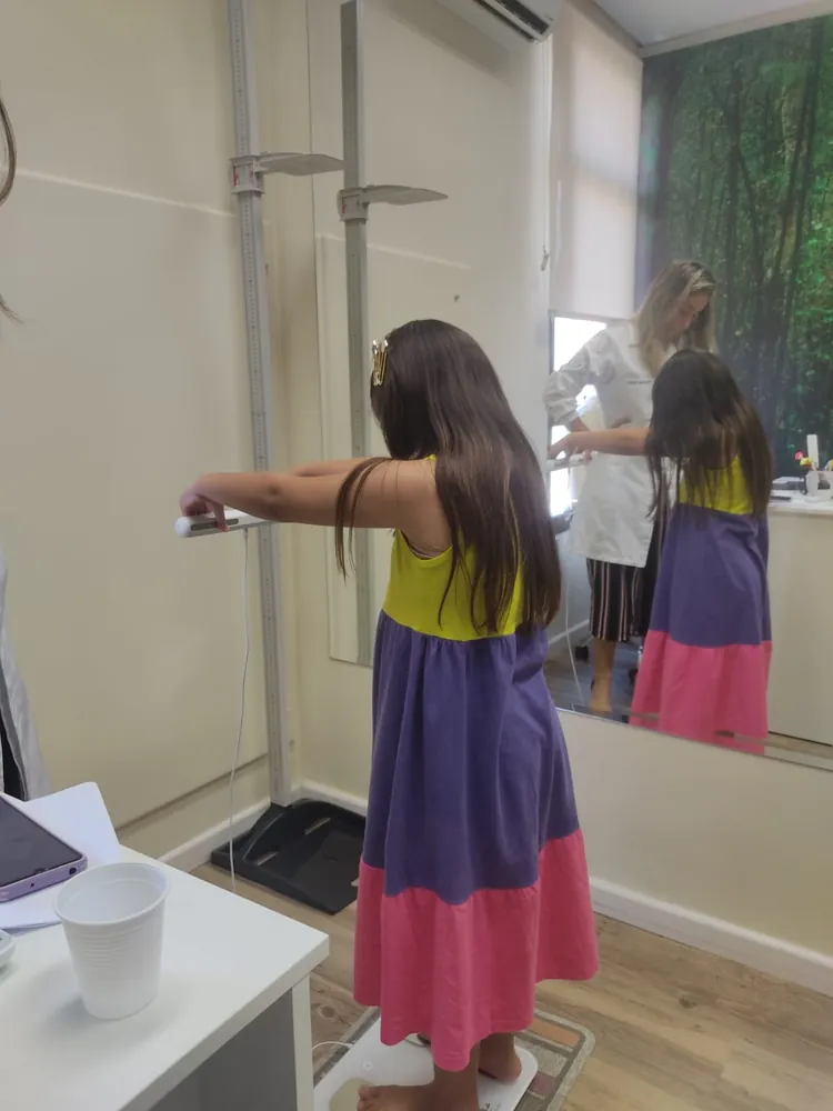 Menina segurando as alças da balança de bioimpedância enquanto Graziella acompanha a avaliação, refletida no espelho