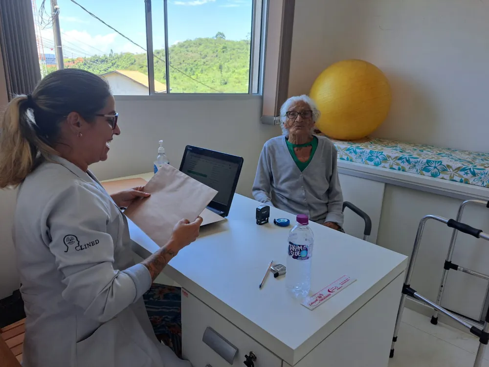 Graziella de jaleco conversando e sorrindo com uma paciente idosa durante a consulta, à mesa do consultório