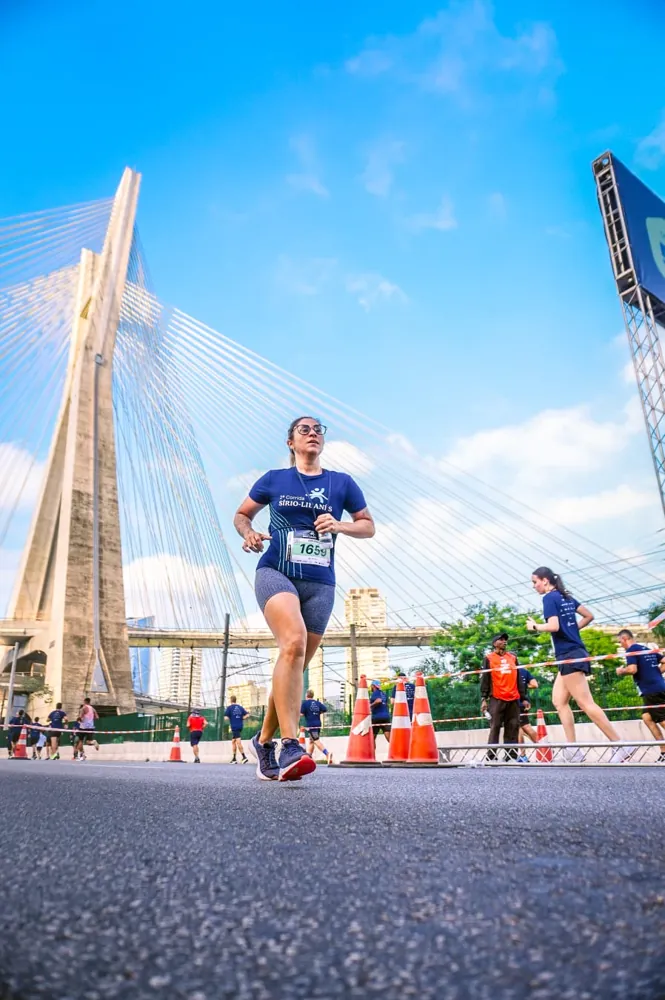 Graziella correndo de camiseta azul e número de peito em uma prova de rua, com a ponte estaiada de São Paulo ao fundo