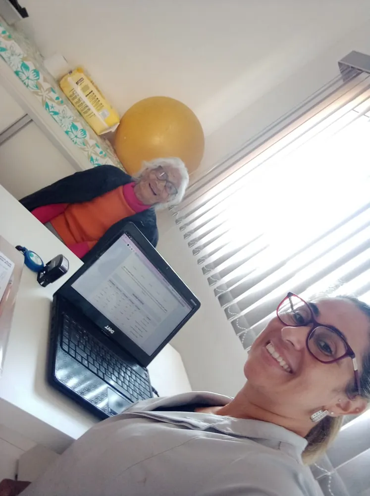 Selfie de Graziella sorrindo no consultório, com uma paciente idosa sorrindo ao fundo