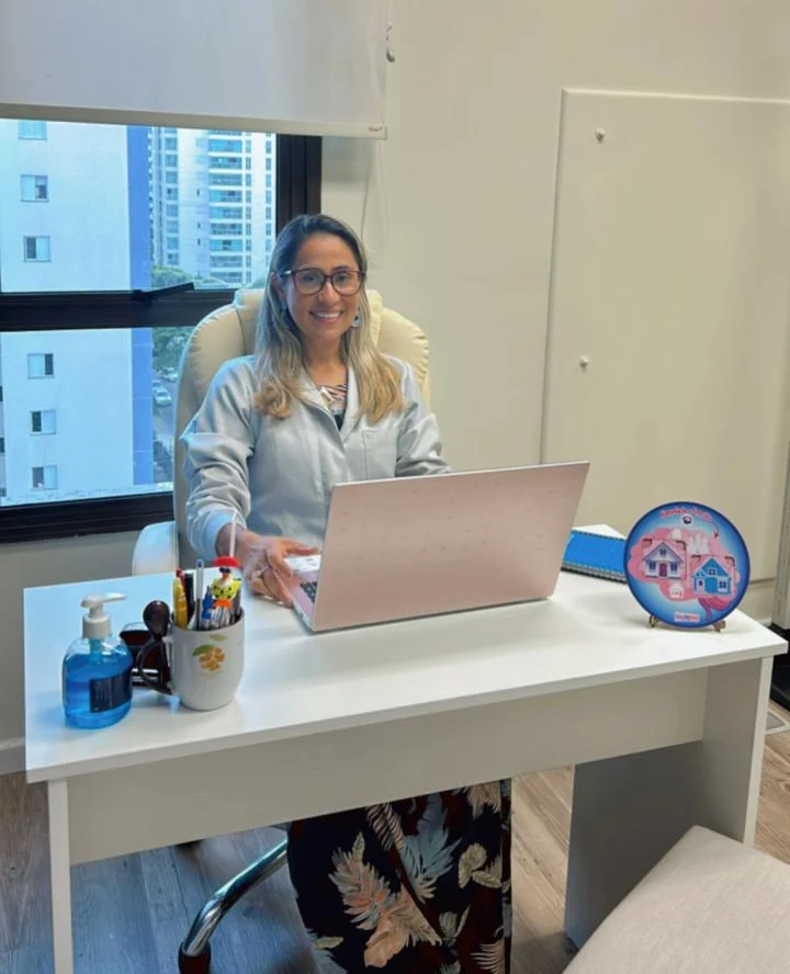 Graziella Santos de óculos e jaleco, sorrindo à mesa do consultório com o notebook aberto